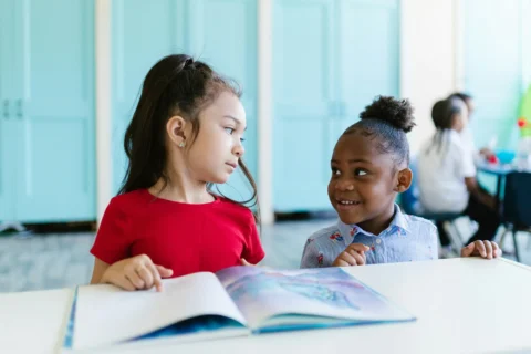 Two young girls sit at a table with an open book, facing each other and smiling.